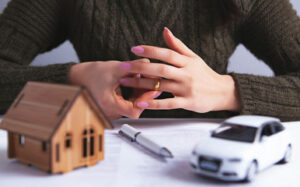 divorce - a woman removing her wedding ring, with toy car and house in front of her and a marriage contract