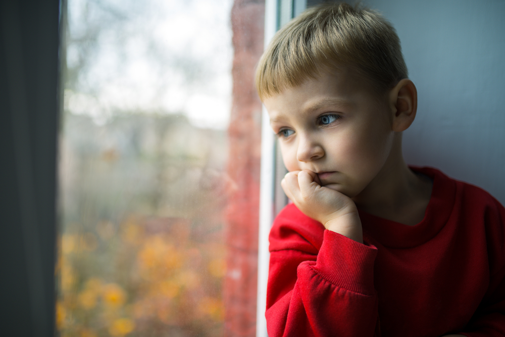 Young boy with blonde hair sitting and looking out the window sadly with his head in his hand.